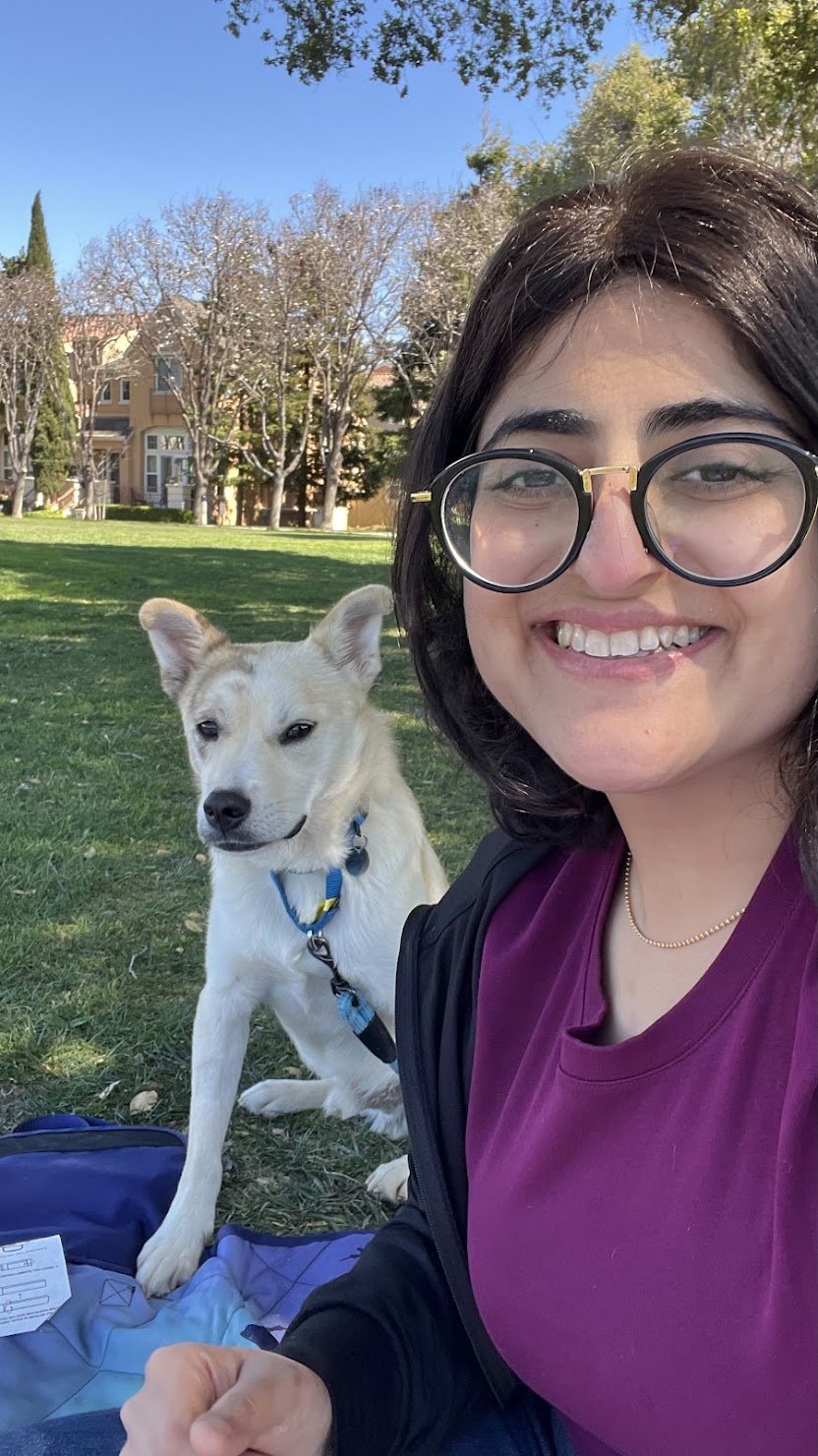 Ghazal sitting in the grass at a park with a light-colored dog